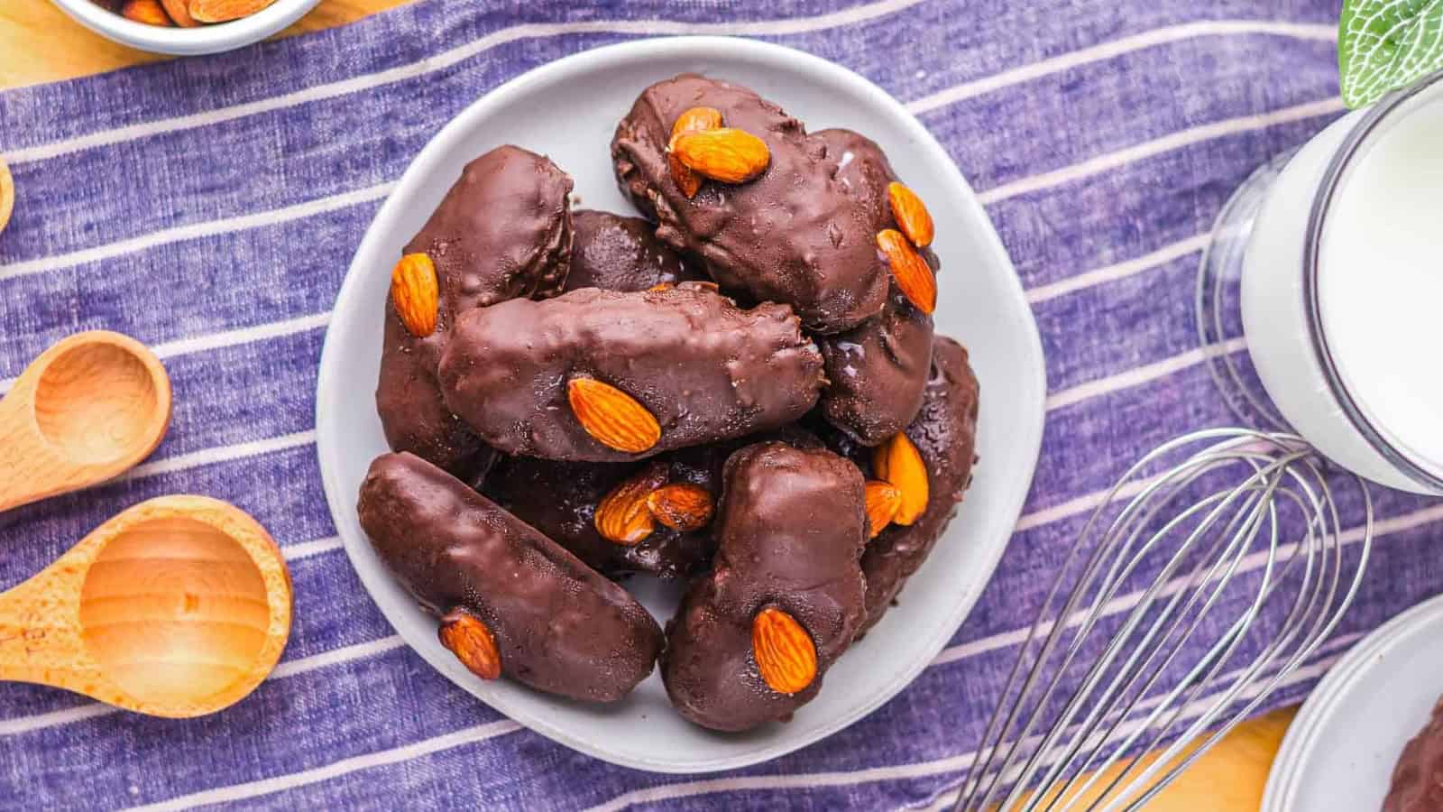 A plate of chocolate-coated bars topped with almonds, placed on a striped cloth next to a whisk.