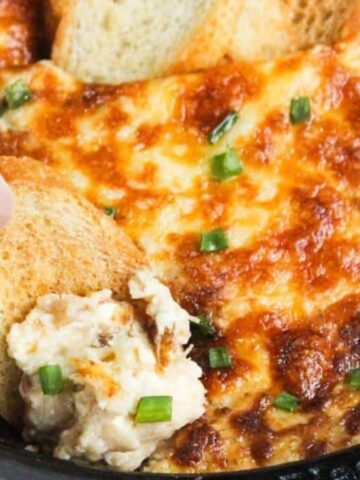 Hand dipping toasted bread into cheesy baked dip topped with chopped green onions in a skillet.
