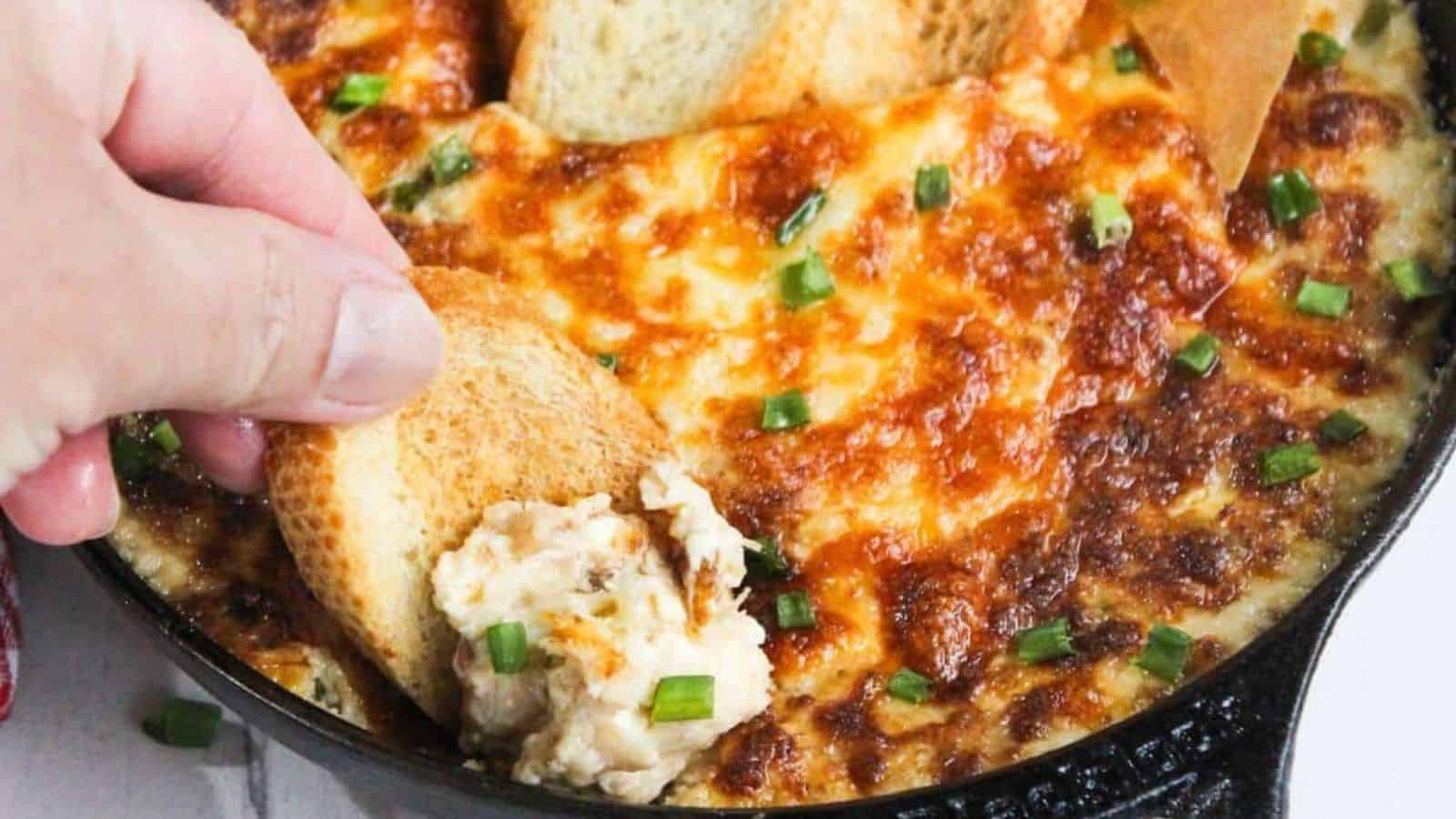 Hand dipping toasted bread into cheesy baked dip topped with chopped green onions in a skillet.