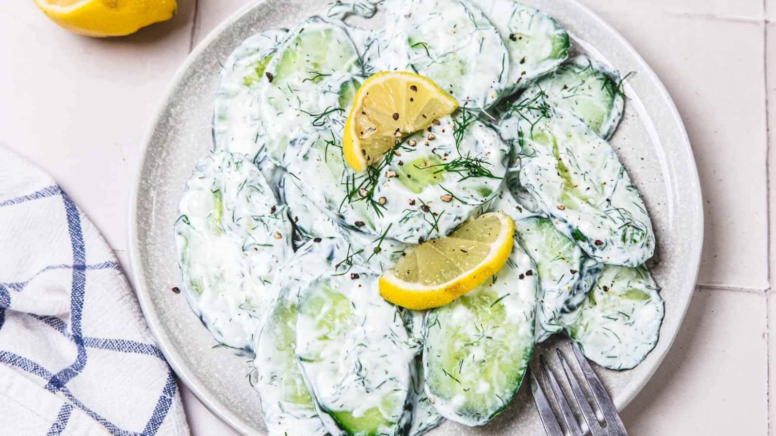 Sliced cucumber salad with creamy dressing, fresh dill, and lemon wedges on a white plate with a fork.