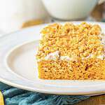 A slice of orange cake with white frosting and chopped nuts on top, served on a white plate.