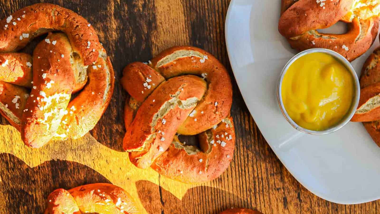 Soft pretzels with coarse salt on a wooden surface, with mustard dipping sauce on a white plate.