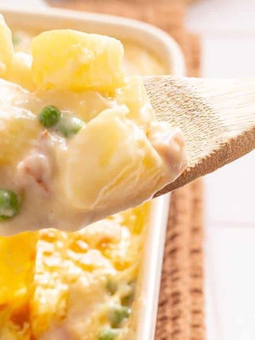 A wooden spoon scoops cheesy casserole with peas and chunks of meat from a baking dish.
