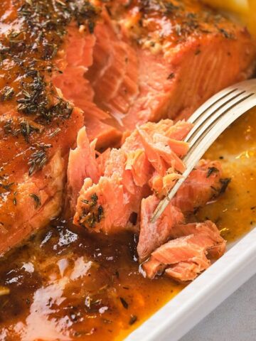 A fork flakes baked salmon with glaze and herbs on a platter, garnished with lemon wedges.