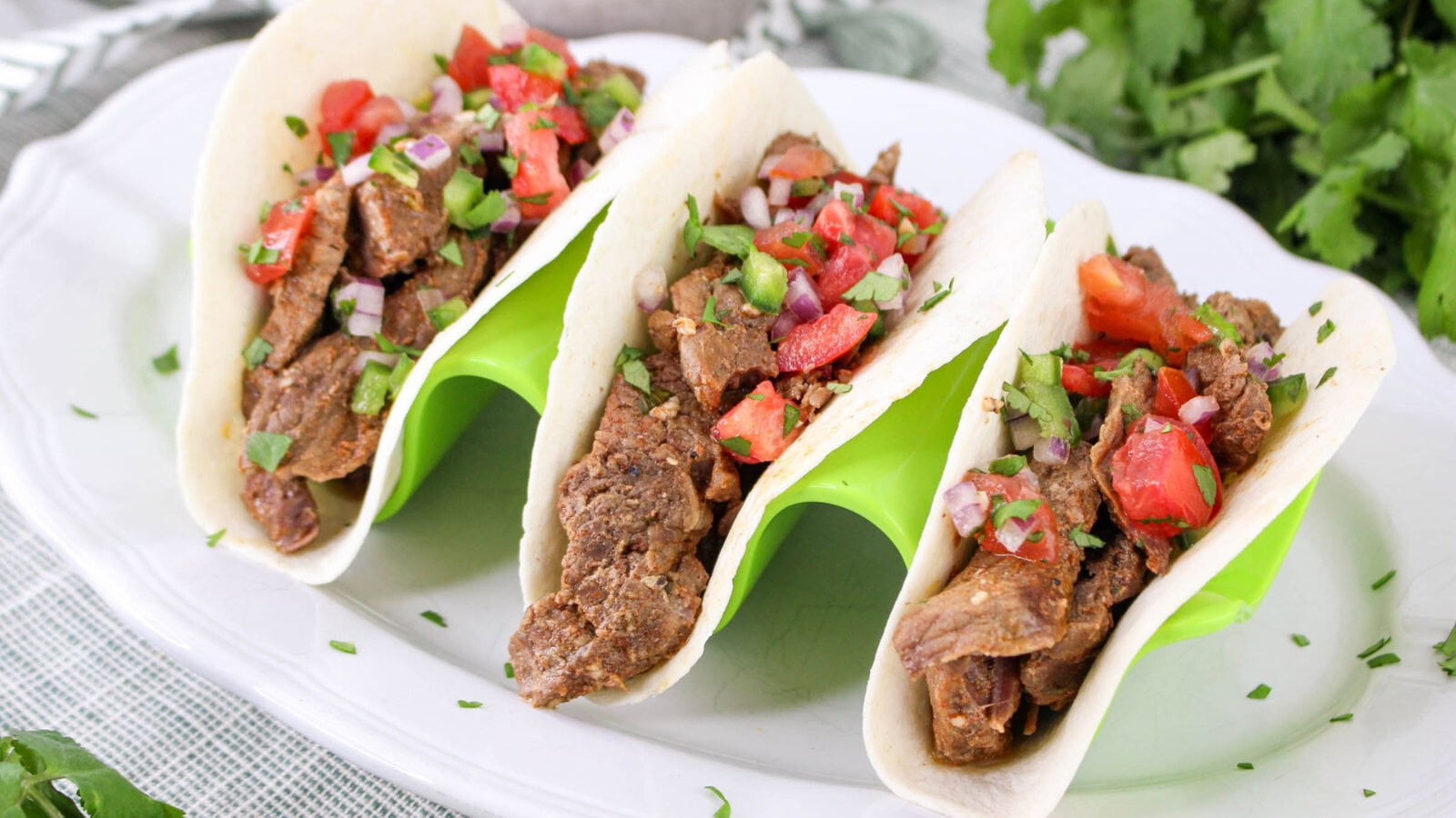Three beef tacos with diced tomatoes, onions, and cilantro served in soft tortillas on a white plate with a green holder.