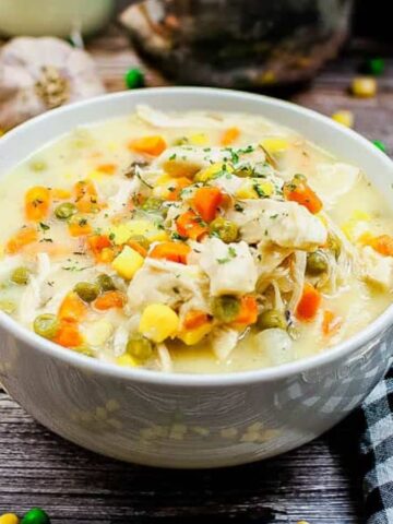 A bowl of creamy chicken and vegetable soup with a spoon and checkered napkin on a wooden table.