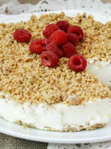 A creamy cheesecake topped with crumbs and fresh raspberries, with a slice missing, on a white plate.