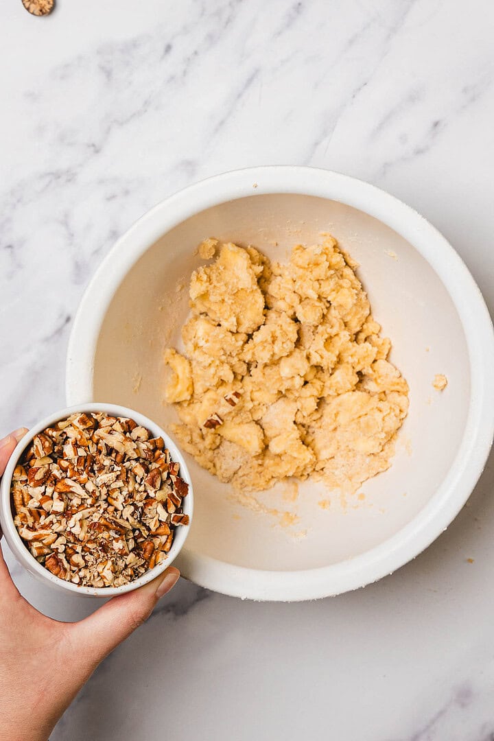 A hand holds a bowl of chopped pecans above a bowl of sweet potato casserole with marshmallows on a marble surface.
