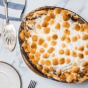 A Sweet Potato Casserole with marshmallows, baked to perfection and topped with golden toasted marshmallows, sits beside empty plates, a fork, and a serving spatula.