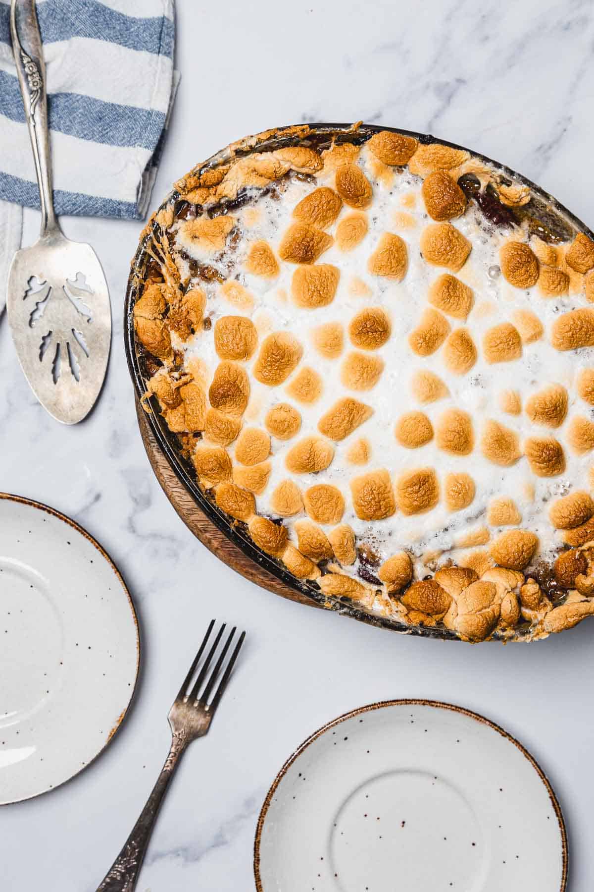 A Sweet Potato Casserole with marshmallows, baked to perfection and topped with golden toasted marshmallows, sits beside empty plates, a fork, and a serving spatula.