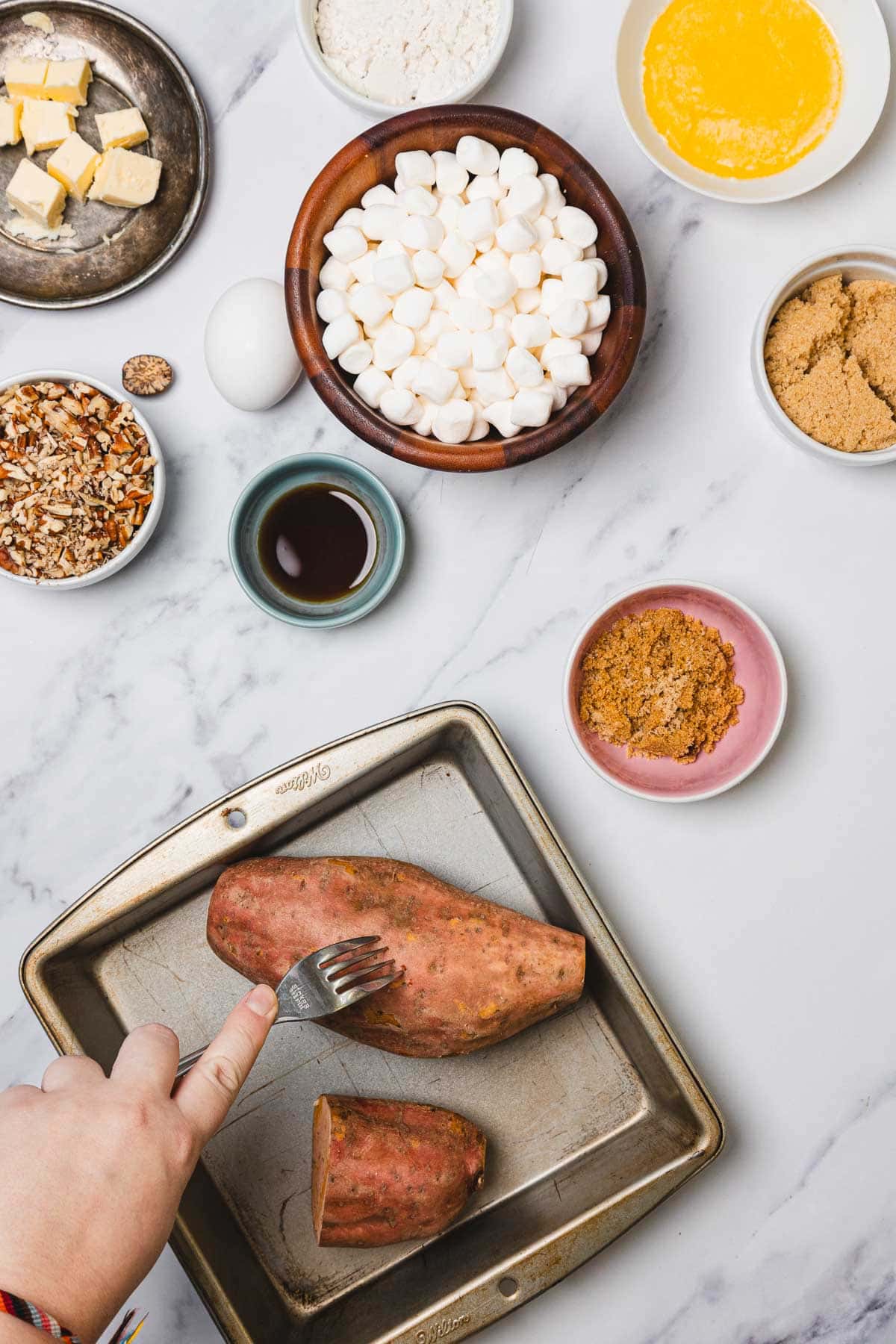 A hand pricks sweet potatoes with a fork on a tray, surrounded by ingredients in bowls—perfect for preparing a classic sweet potato casserole with marshmallows on a marble counter.