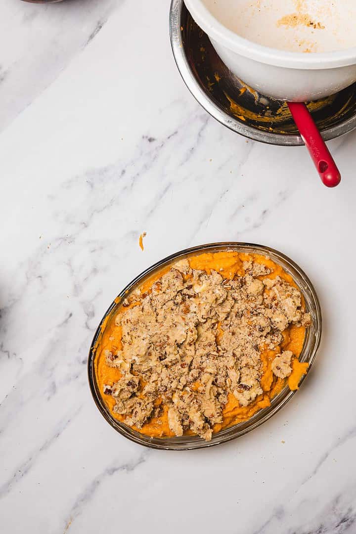An oval dish of Sweet Potato Casserole with pecan topping sits on a marble counter, mixing bowls above—ideal for trying your favorite Sweet Potato Casserole Recipe, with or without marshmallows.