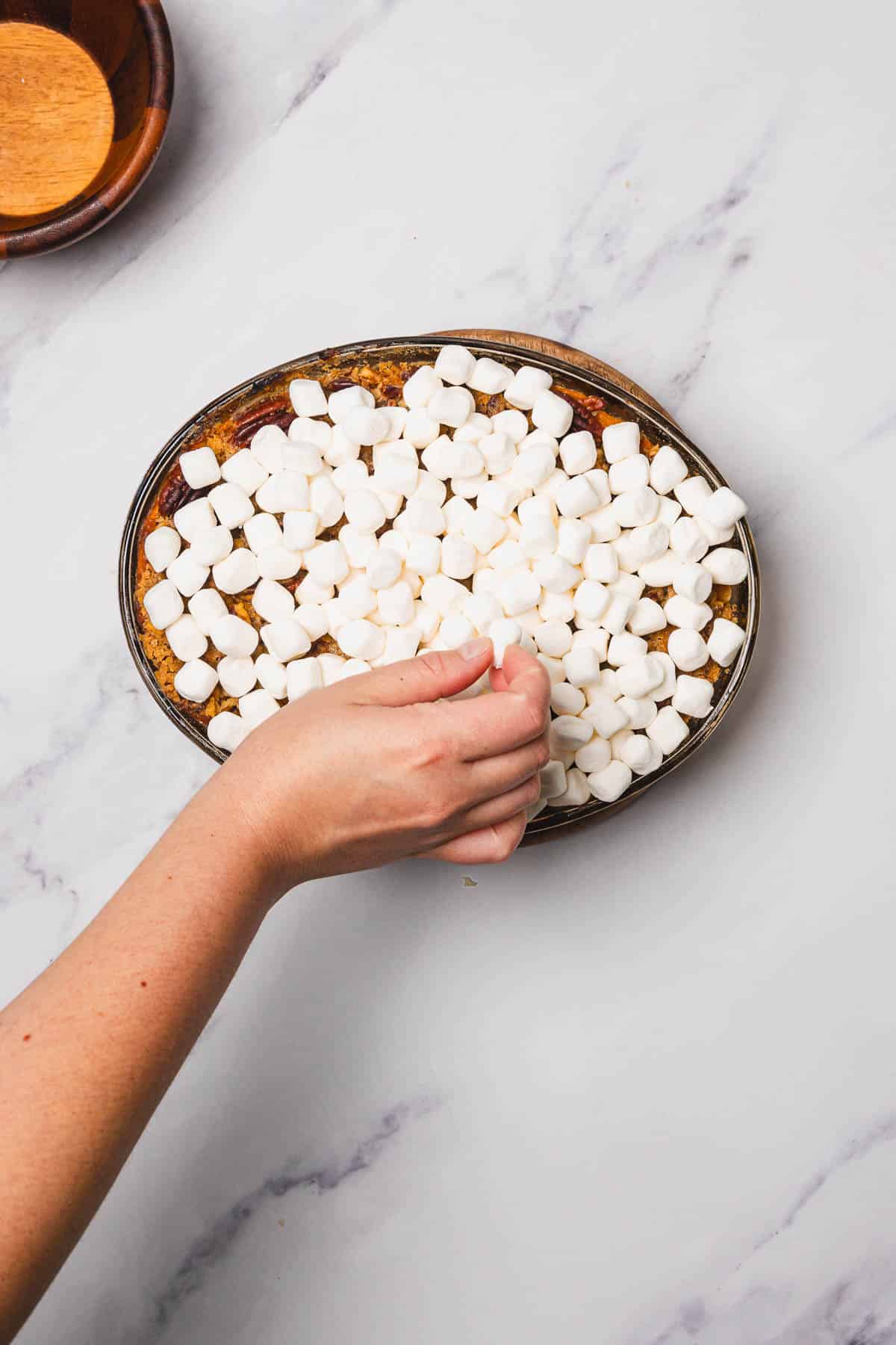 A hand places mini marshmallows on top of a sweet potato casserole in an oval dish on a marble surface.