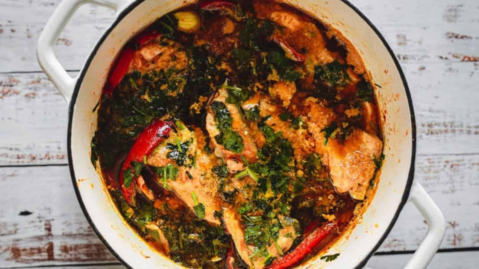 A pot of fish stew with herbs, red peppers, and a rich, colorful broth on a white wooden surface.
