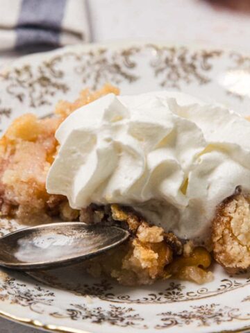 A serving of peach cobbler topped with whipped cream on a decorative plate with a spoon.
