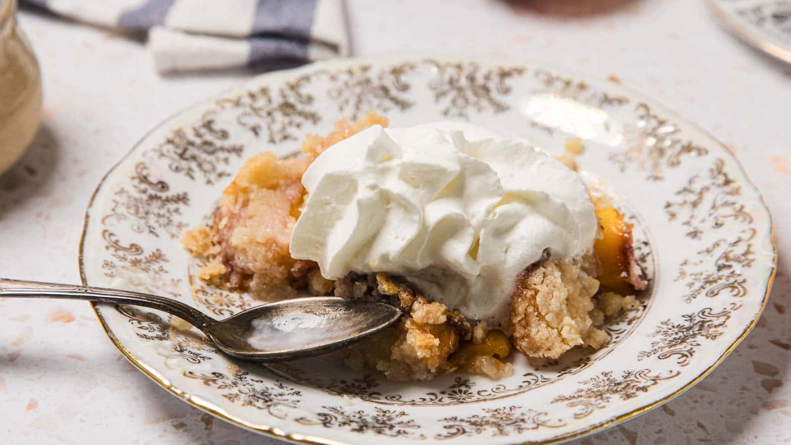 A serving of peach cobbler topped with whipped cream on a decorative plate with a spoon.