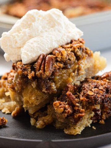 A slice of pecan pie topped with whipped cream on a black plate, with a baking dish in the background.