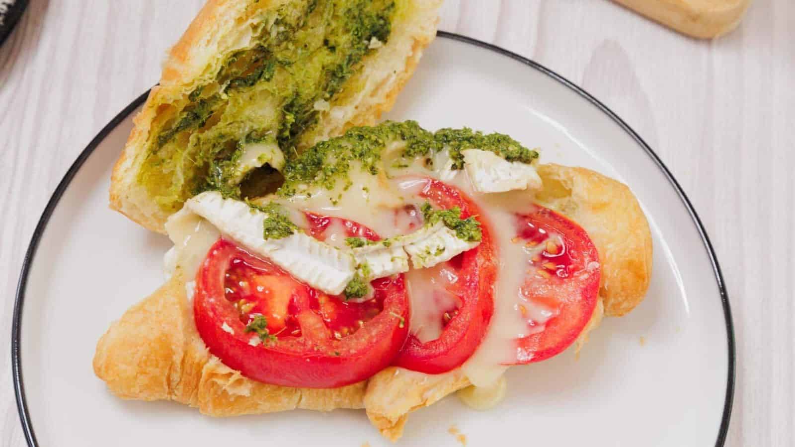 A croissant sandwich with tomato slices, brie cheese, and pesto on a white plate.