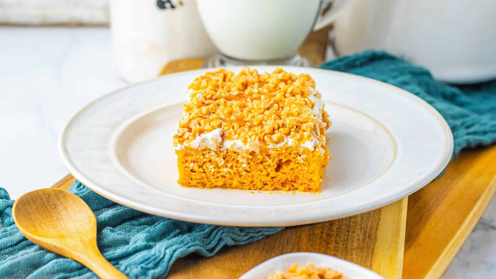 A slice of pumpkin cake with frosting and chopped nuts on a white plate, next to a wooden spoon.