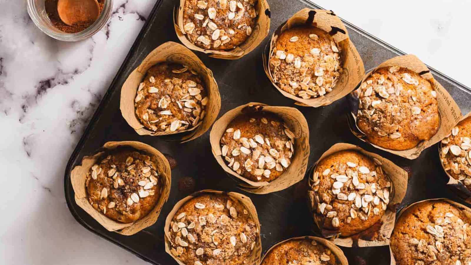 A muffin topped with oats is placed on a decorative paper wrapper. The muffin sits on a marble surface, with crumbs scattered around it.