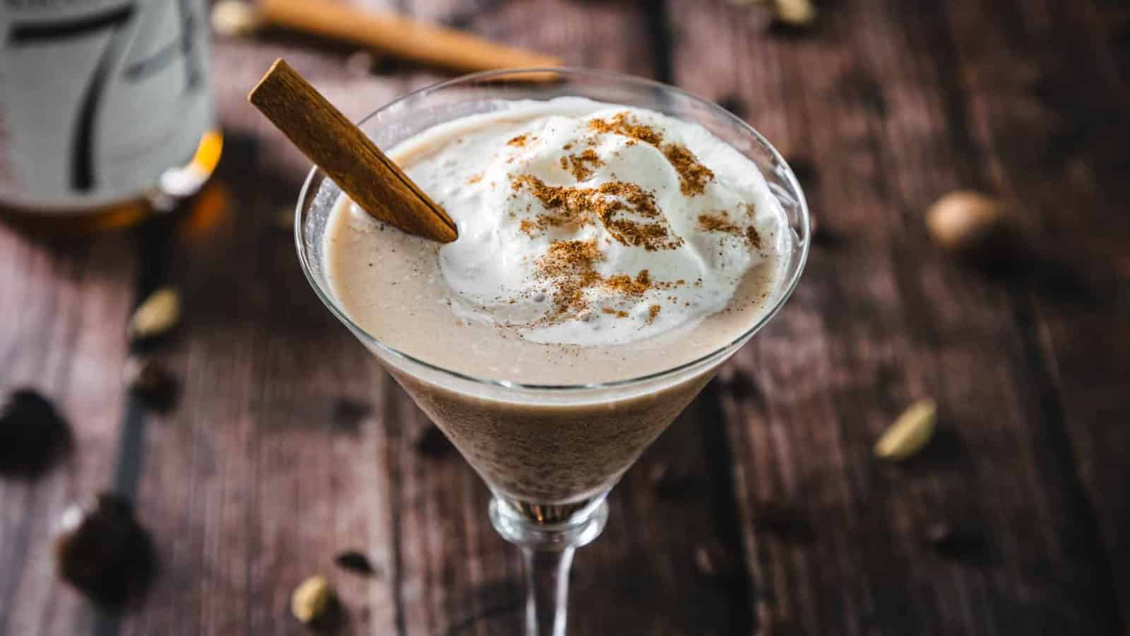 A creamy cocktail in a martini glass topped with whipped cream, cinnamon, and a cinnamon stick.