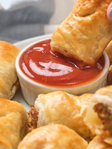A hand dips a puff pastry sausage roll into a bowl of ketchup, surrounded by more sausage rolls.