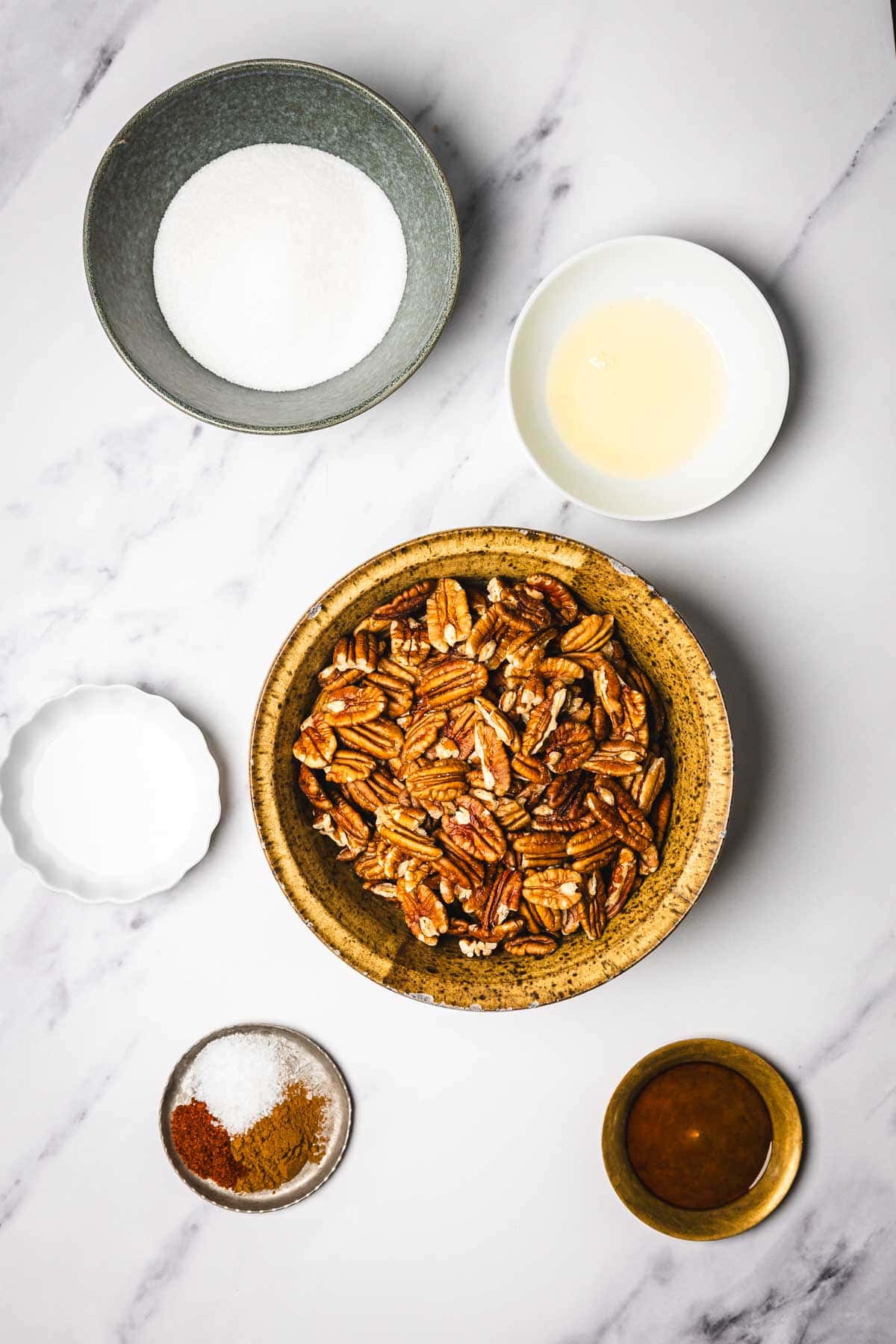 Bowls with ingredients for slow cooker candied pecans&mdash;pecans, sugar, oil, spices, and syrup&mdash;arranged on a white marble surface seen from above.
