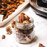 A glass jar filled with slow cooker candied pecans, with more pecans scattered on a white surface nearby.