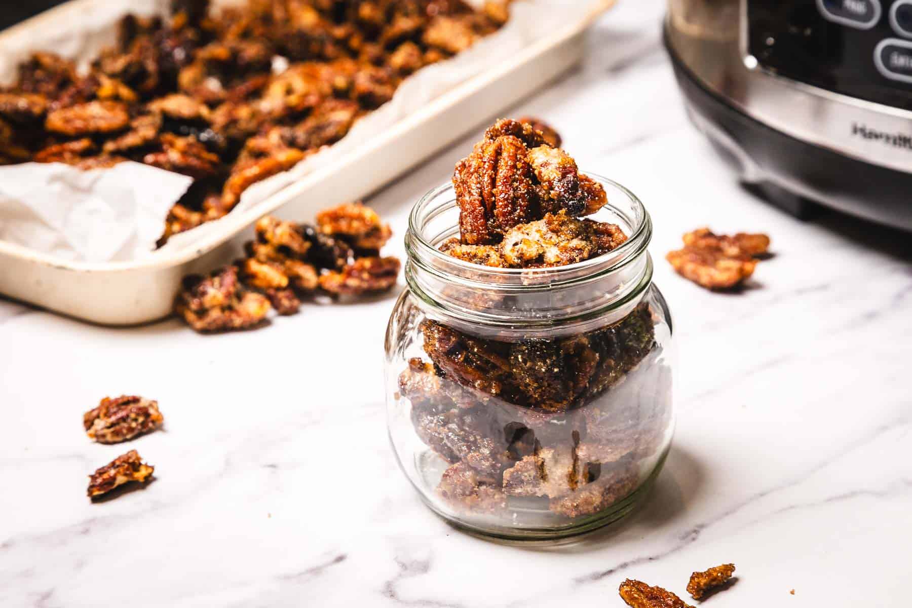 A glass jar filled with slow cooker candied pecans sits on a marble counter, with more pecans in a tray behind it.