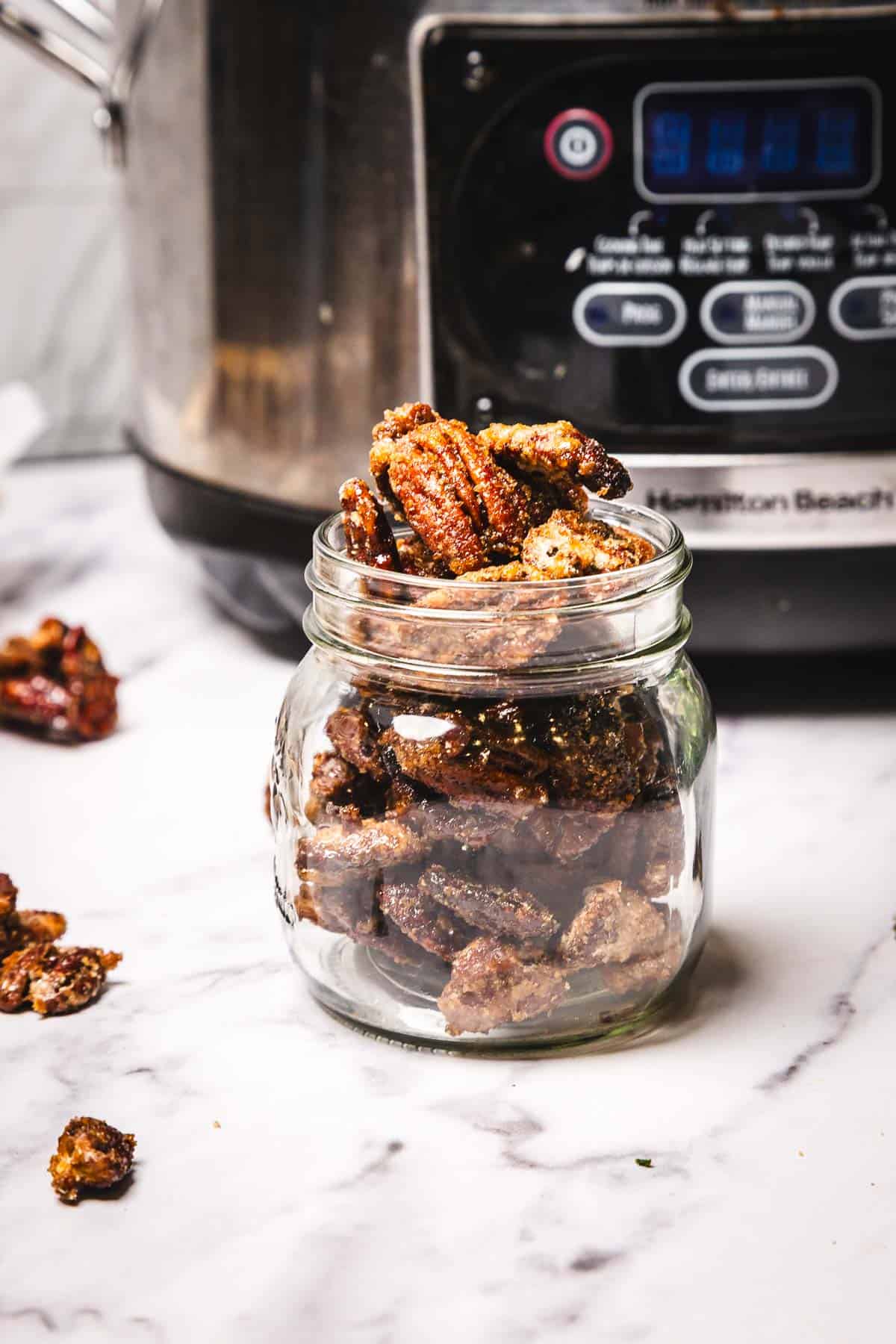 A glass jar filled with slow cooker candied pecans sits on a marble counter near the slow cooker.