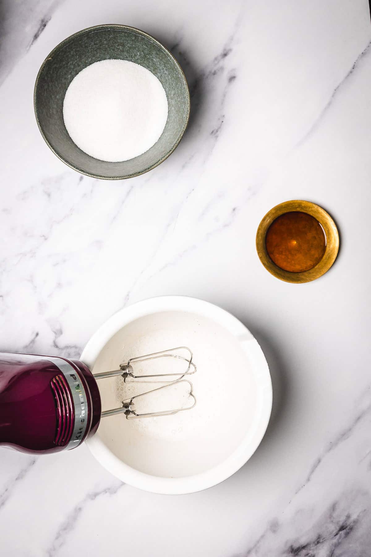 A hand mixer in a white bowl, with a bowl of sugar and a small dish of vanilla extract nearby on a marble surface&mdash;perfect for prepping slow cooker candied pecans.