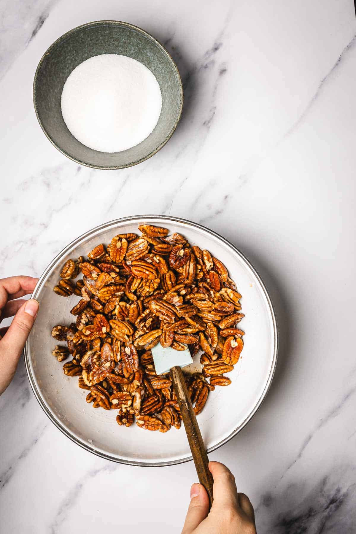 Hands mixing slow cooker candied pecans in a bowl with a spatula; a bowl of sugar sits nearby on a marble surface.
