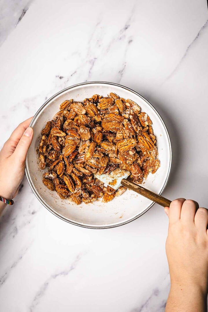 Hands mixing slow cooker candied pecans in a white bowl with a spoon on a marble surface.