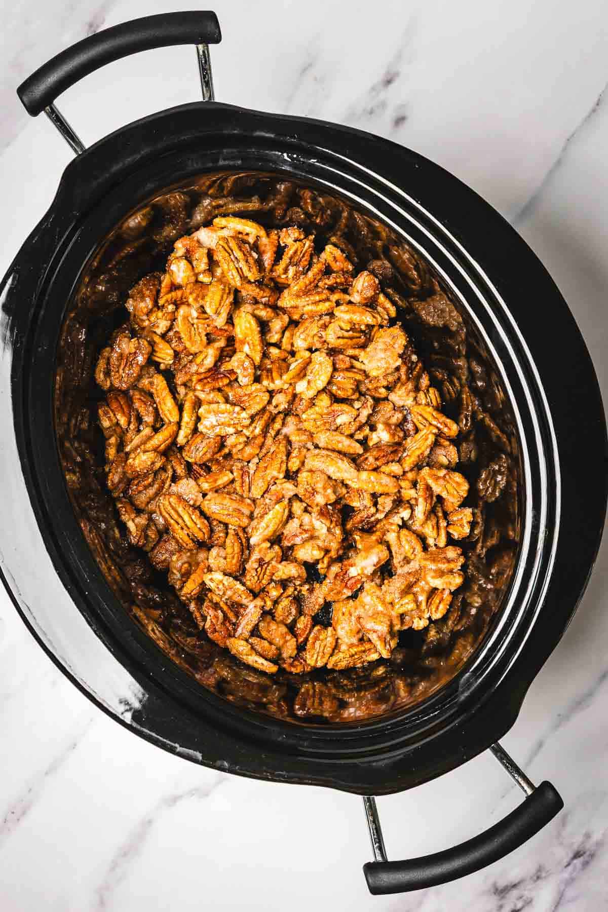 Top view of slow cooker candied pecans in a black slow cooker on a white marble surface.