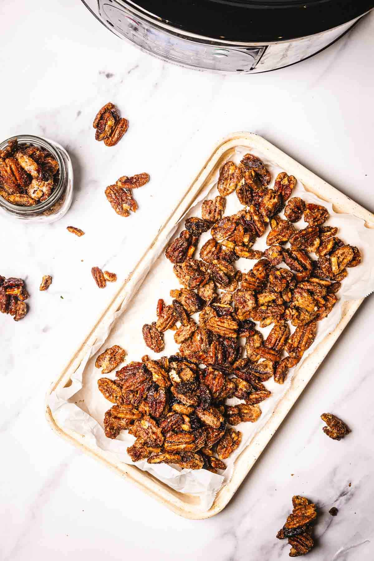 A tray of slow cooker candied pecans on parchment paper, with a jar and scattered pecans on a white surface.