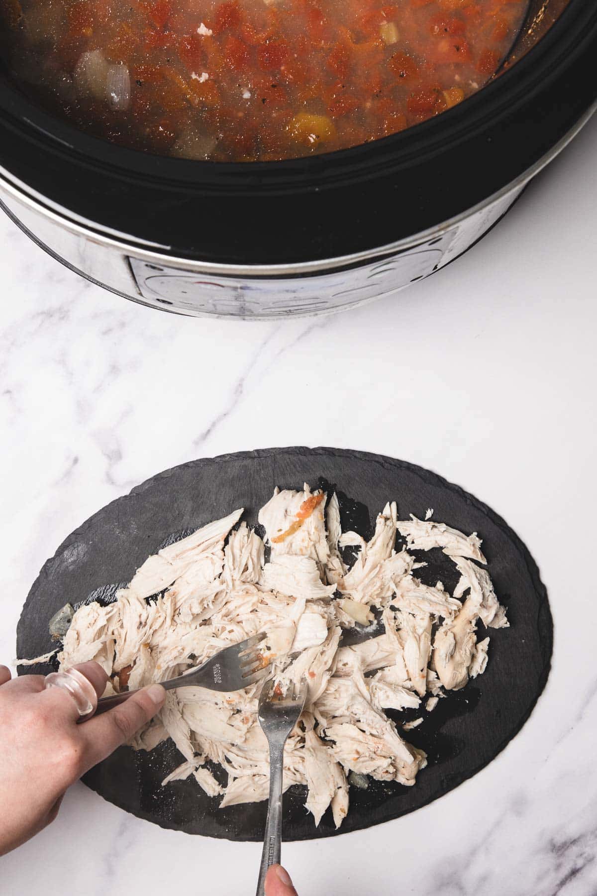 Two hands shred cooked chicken with forks on a black slate board next to a slow cooker, perfect for preparing hearty Chicken Spaghetti or soup.