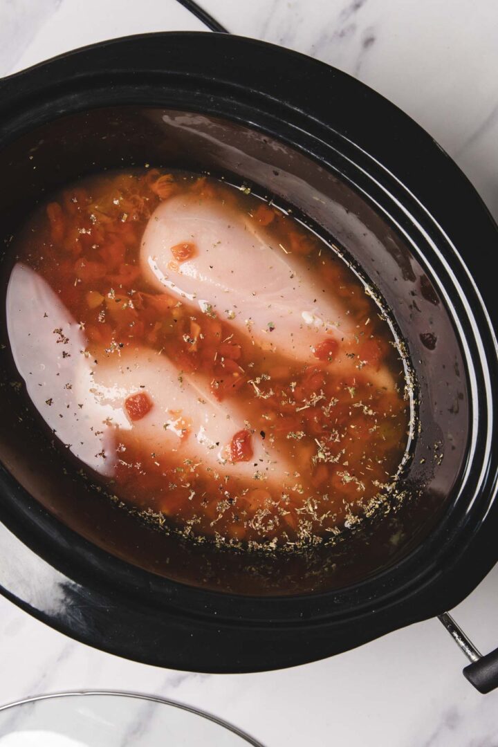 Slow Cooker Chicken breasts sit in a slow cooker topped with tomato sauce and herbs, perfect for an easy base for delicious Chicken Spaghetti.
