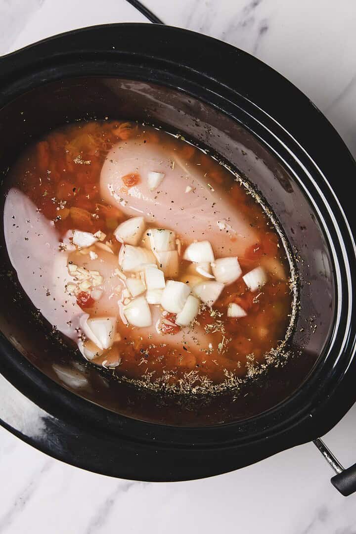 Raw chicken breasts, chopped onions, herbs, and broth in a slow cooker&mdash;perfect for making flavorful Slow Cooker Chicken Spaghetti.