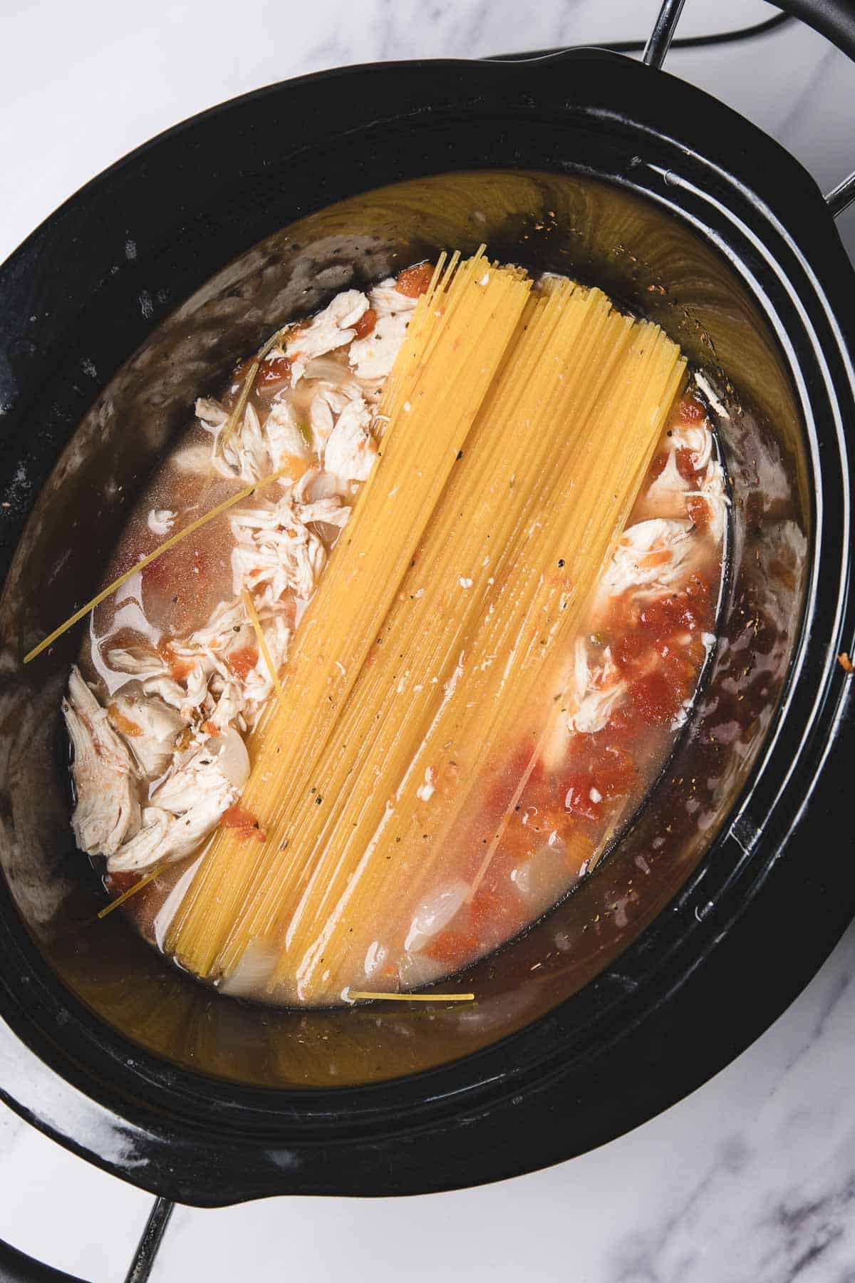 Uncooked spaghetti and shredded chicken in broth inside a slow cooker, ready to become a delicious chicken spaghetti meal.