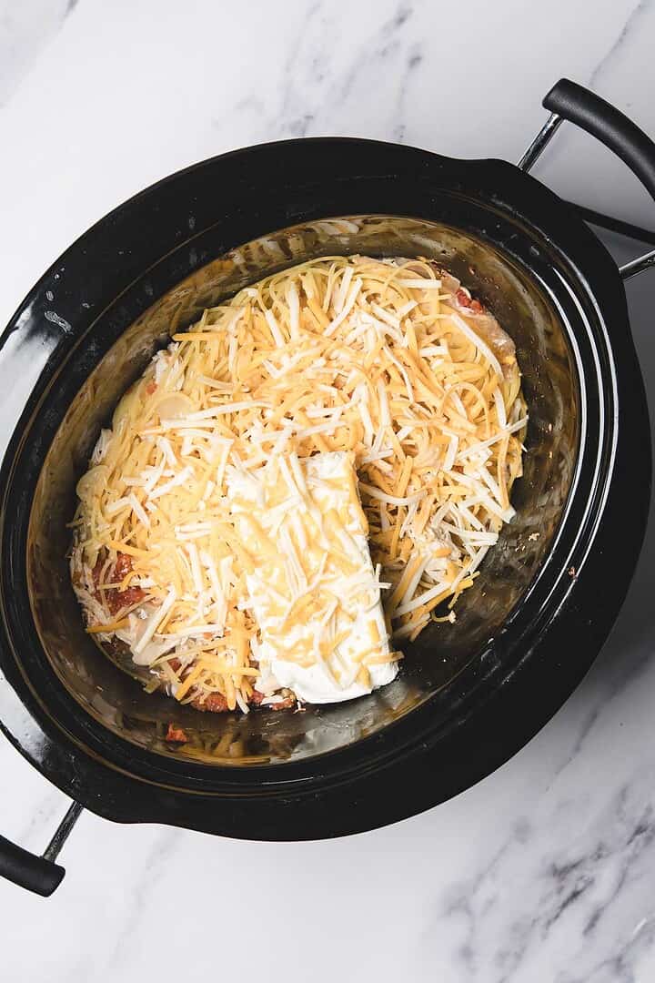 A slow cooker filled with shredded cheese and a block of cream cheese sits atop tender Slow Cooker Chicken, making the perfect base for creamy Chicken Spaghetti.