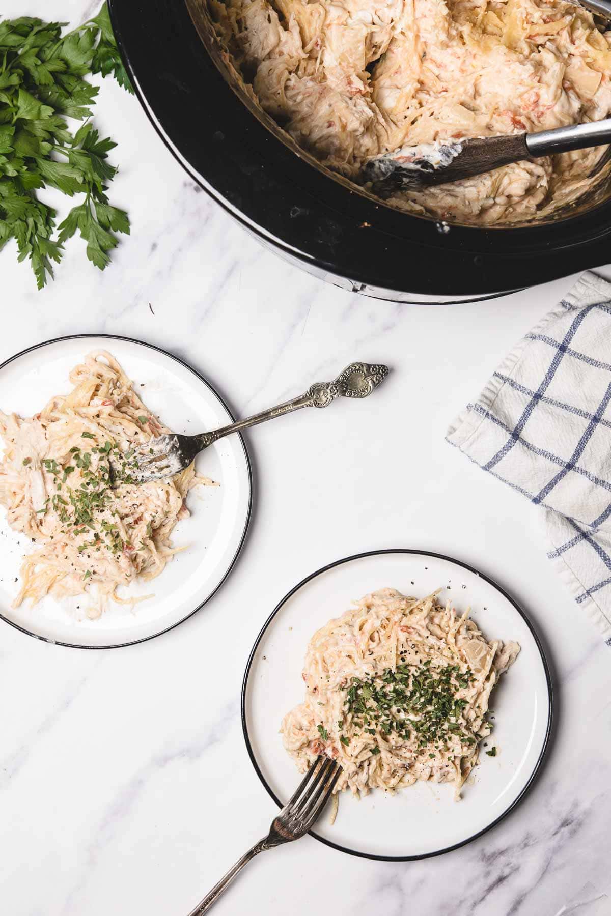Two plates of creamy chicken spaghetti with herbs sit beside a slow cooker, fresh parsley, and a striped towel&mdash;an easy chicken spaghetti recipe for your next cozy meal.