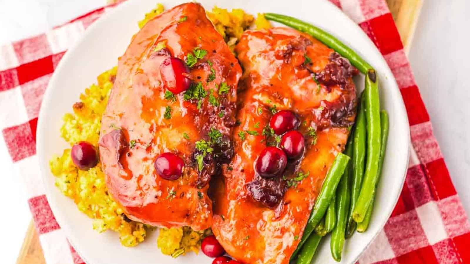 Two glazed chicken breasts with cranberries, green beans, and rice on a white plate with a red checkered napkin.
