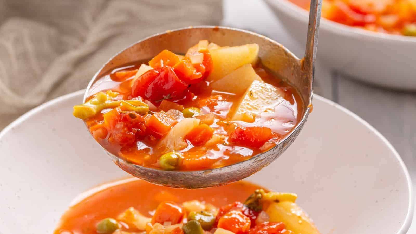 A ladle serving vegetable soup with tomatoes, potatoes, and green peas into a white bowl, with a blurred bowl in the background.