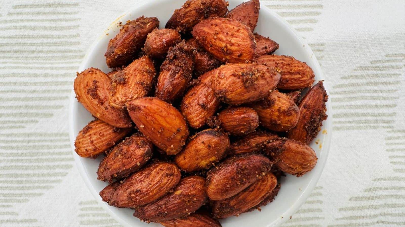 A white bowl filled with roasted, seasoned almonds is placed on a white and grey striped fabric.