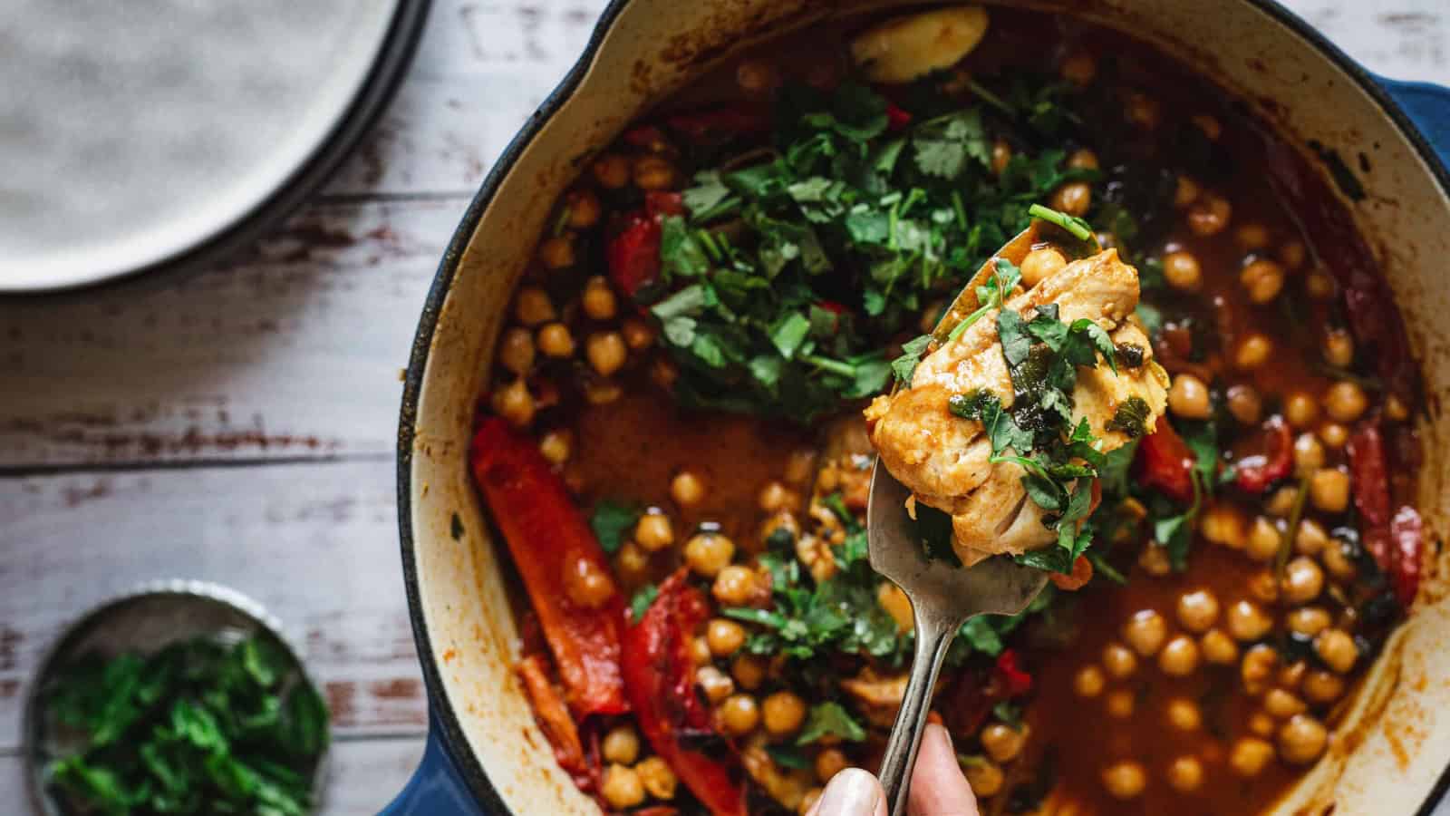 A fork holds chickpea stew with herbs over a pot filled with stew, chickpeas, and chopped greens.