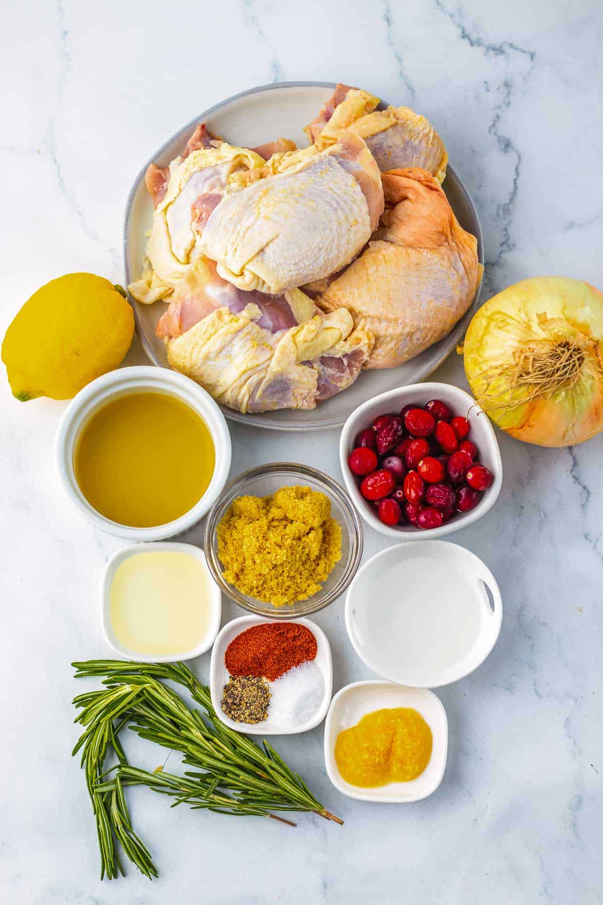 Raw turkey pieces, lemon, onion, cranberries, oil, spices, broth, and herbs arranged on a white surface—perfect ingredients for cranberry chicken or roasted chicken thighs recipes.