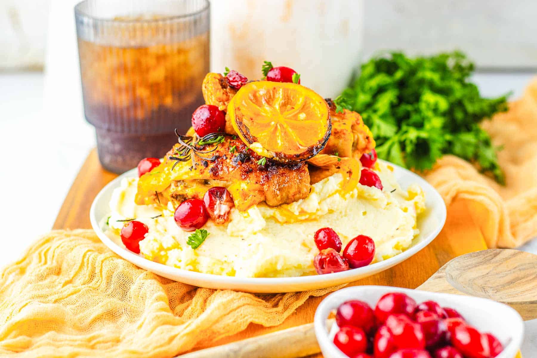 Grilled cranberry chicken, with lemon and herbs, served over mashed potatoes and topped with cranberries. Enjoy this roasted cranberry chicken thighs dish with a refreshing drink and fresh greens in the background.