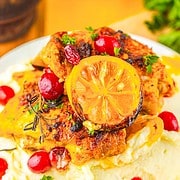 Roasted Cranberry Chicken Thighs with lemon slices, cranberries, and herbs rest atop creamy mashed potatoes, served on a white plate.