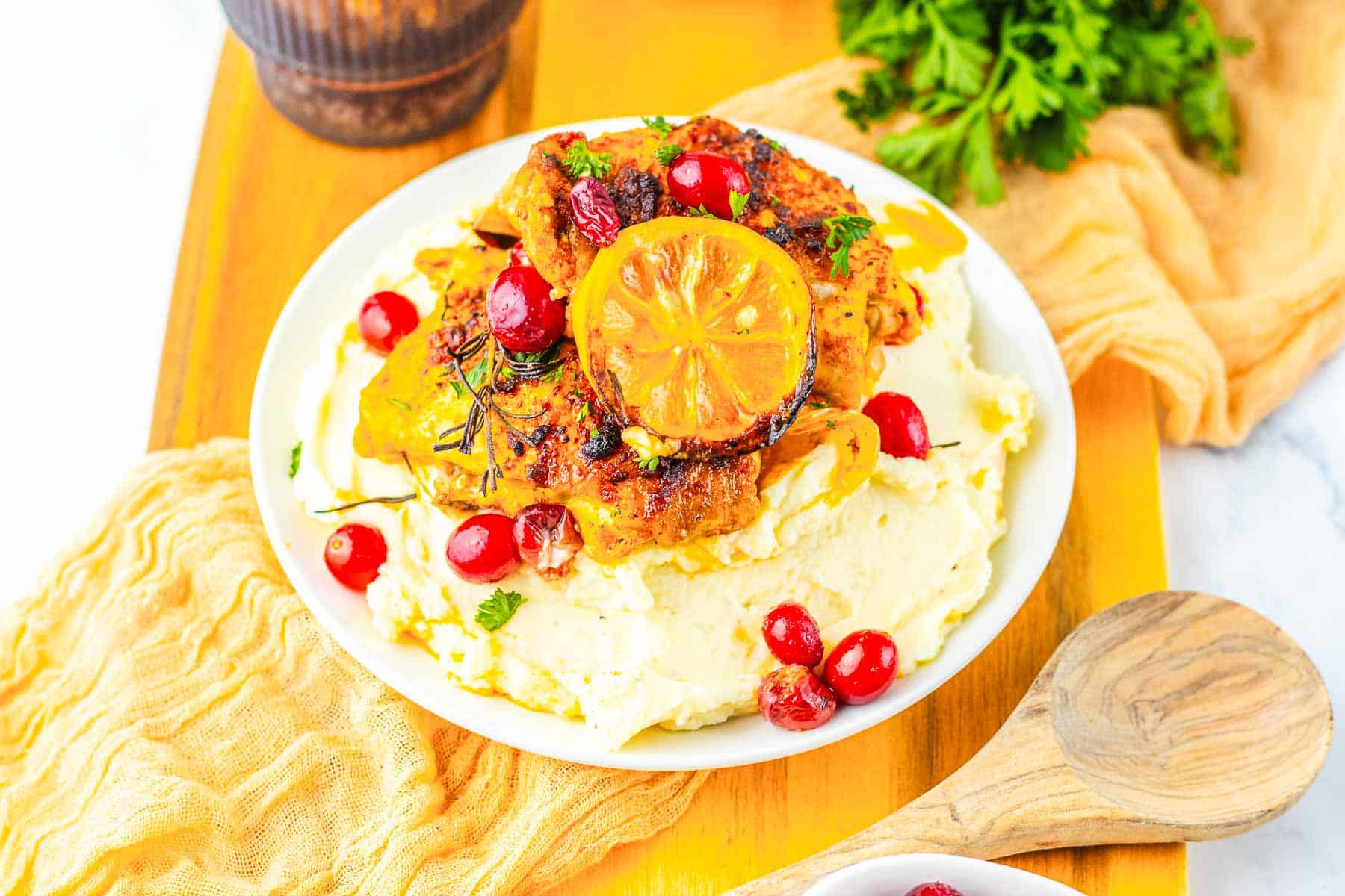 Plate of roasted cranberry chicken on mashed potatoes, topped with a lemon slice and cranberries, served on a wooden board.