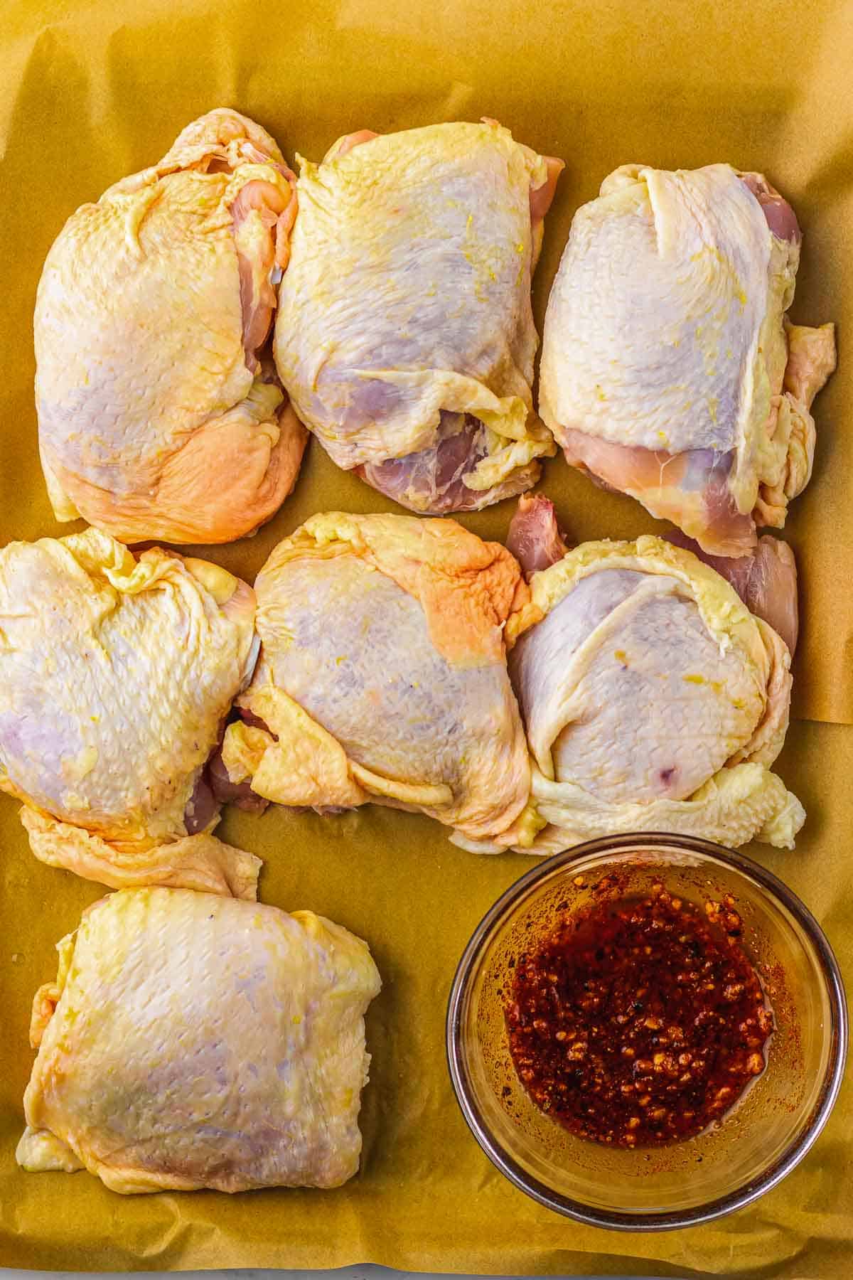 Six raw chicken thighs on parchment paper next to a small bowl of dark, seasoned marinade, ready to become delicious Roasted Cranberry Chicken Thighs.